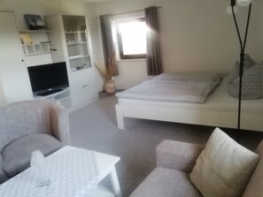 A room with a sofa, a small table, a bed, a TV, and a window with curtains.