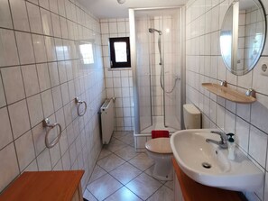 Ein Badezimmer mit weißen Fliesen, einer Dusche, einer Toilette, einem Waschbecken und einem Spiegel.