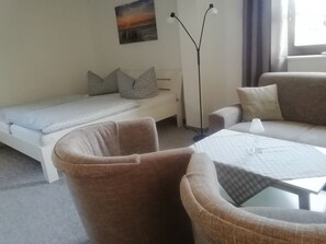 A small, cozy room with a single bed, a sofa, two armchairs, a table, and a floor lamp.