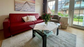 A living room with a red sofa, a glass coffee table, a vase with flowers, and a painting of a beach scene. - 005 - Hooge (BC.n3) - BUE - Apartment house Seewind (Büsum)