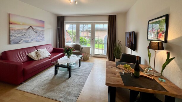 A living room with a red sofa, a coffee table, a TV, and a large window with curtains. - 005 - Hooge (BC.n3) - BUE - Apartment house Seewind (Büsum)