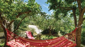 Un hamac est suspendu entre deux arbres dans un jardin, avec une structure en pierre en arrière-plan.