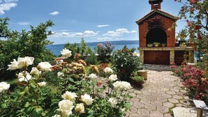Une construction en briques, dotée d’une entrée en arc de cercle, entourée d’un jardin où poussent différentes espèces de fleurs et offrant une vue sur la mer.