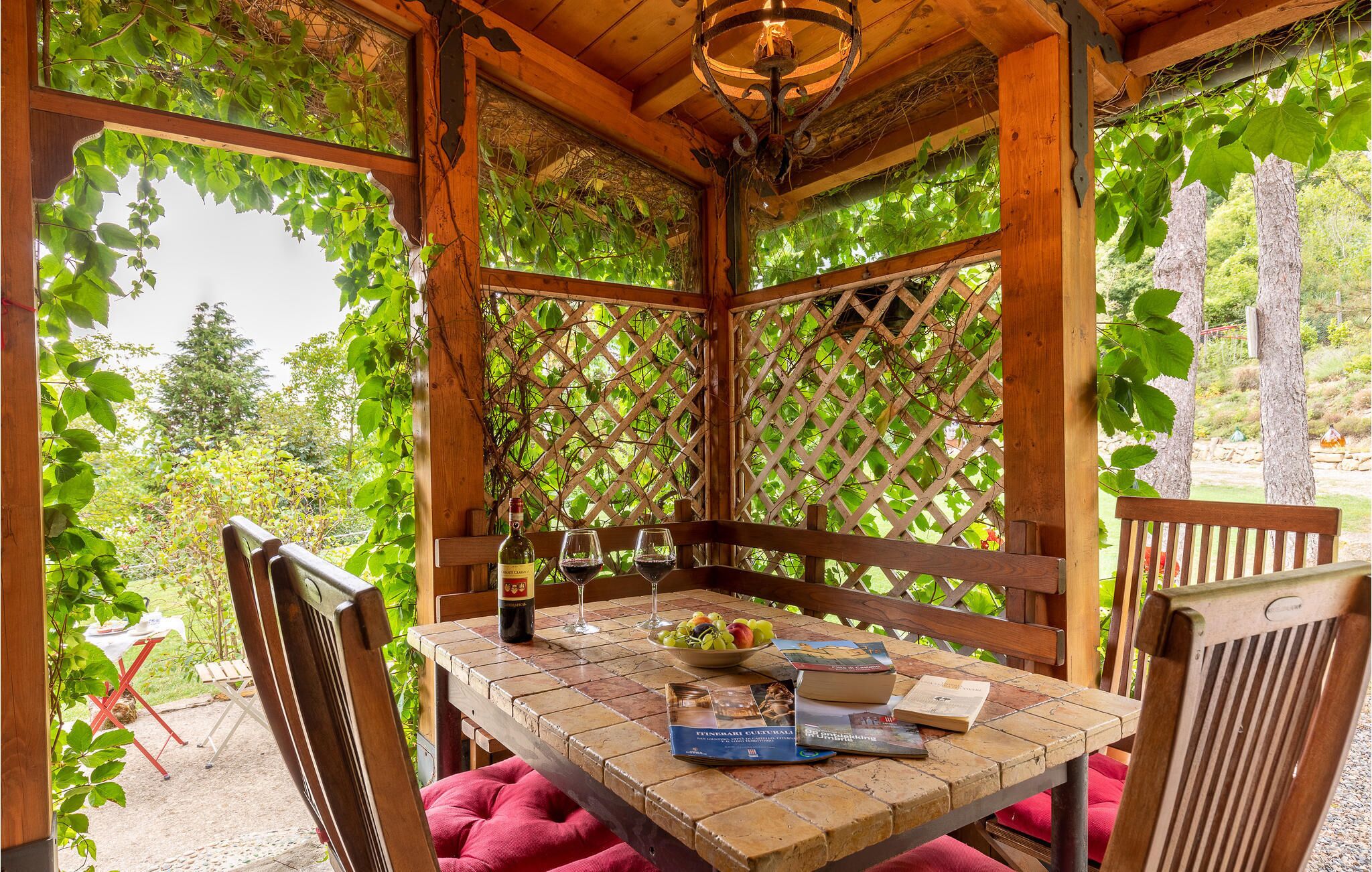 Un gazebo in legno con un tavolo apparecchiato per due persone, con una bottiglia di vino, bicchieri e una ciotola di frutta.