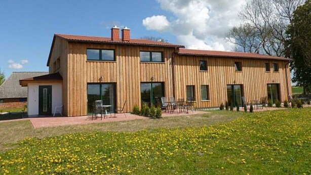 Een houten huis met twee verdiepingen, een rood dak, grote ramen en een klein terras met tuinmeubilair.