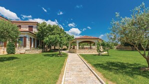 Ein gepflasterter Weg führt zu einem überdachten Außenbereich mit Ziegeldach, umgeben von gepflegtem Rasen und Bäumen.