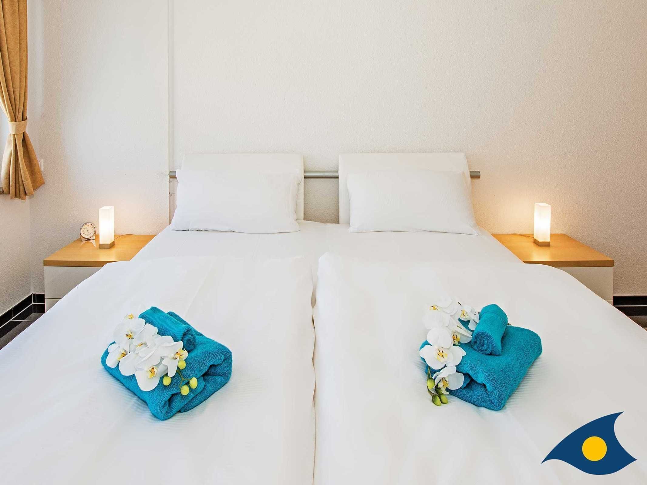 Ein Hotelzimmer mit zwei Betten, weißen Handtüchern und Blumen. An den Nachttischen stehen Lampen und eine Uhr.