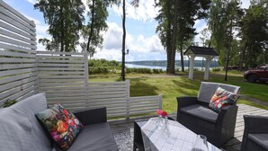 Un área de estar al aire libre con una pérgola blanca, una mesa con mantel a cuadros y vistas a un lago y árboles.