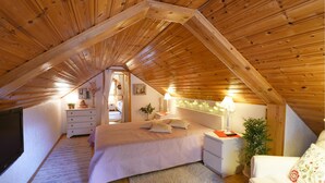 Un dormitorio acogedor en el ático, con techo inclinado, una cama con almohadas, una mesita de noche con lámpara, un aparador y una planta pequeña.
