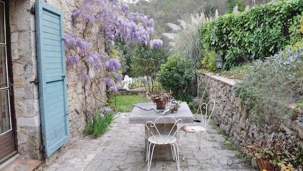 A stone building with a blue shutter, a garden with purple flowers, and two white metal chairs on a stone pathway. - Awesome home in La Roquette sur Siagne (La Roquette sur Siagne)
