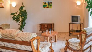 Ein Wohnzimmer mit einer Couch, Sesseln, einem Holzschrank, einem kleinen Tisch mit Flasche und Gläsern sowie einem Fernseher auf einem Schrank.