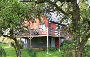 Une maison en bois rouge avec un balcon, entourée d’arbres et d’une cour verdoyante.