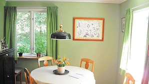 Uma sala de jantar com uma mesa redonda, cadeiras, um candeeiro pendente, uma impressão botânica emoldurada e uma janela com cortinas.