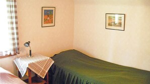 Um quarto pequeno e modesto, com uma cama de solteiro, uma mesa com toalha xadrez, um abajur e duas fotos emolduradas na parede.