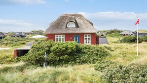 Ein traditionelles rotes Haus mit einem Strohdach, umgeben von Grün und weiteren Häusern in ländlicher Umgebung.