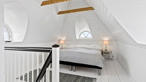 Ein Schlafzimmer mit schrägen Wänden, einem Bett mit gemustertem Überwurf, zwei Nachttischlampen und einem Fenster mit Blick.
