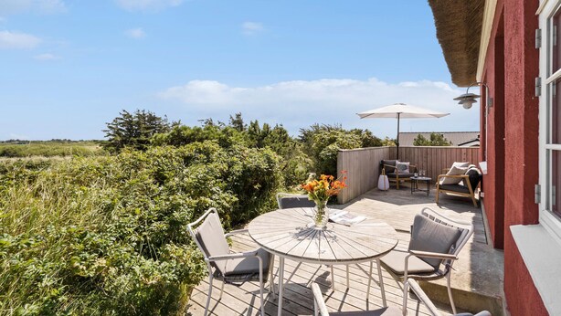 Eine Holzterrasse mit rundem Tisch und Stühlen, ein kleiner Tisch mit Blumen in einer Vase und ein Sonnenschirm.
