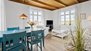 Ein modernes Wohnzimmer mit Holzbalkendecke, weißen Wänden und großen Fenstern. Es gibt einen Esstisch mit Stühlen, eine Couch, einen Fernseher und einen Couchtisch.
