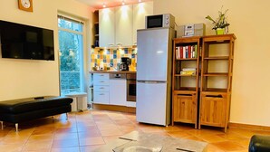 Eine moderne Küche mit Kühlschrank, Mikrowelle und Kaffeemaschine. Ein Holzregal mit Büchern und einer Topfpflanze ist vorhanden. Im Wohnbereich befindet sich eine schwarze Lederbank.