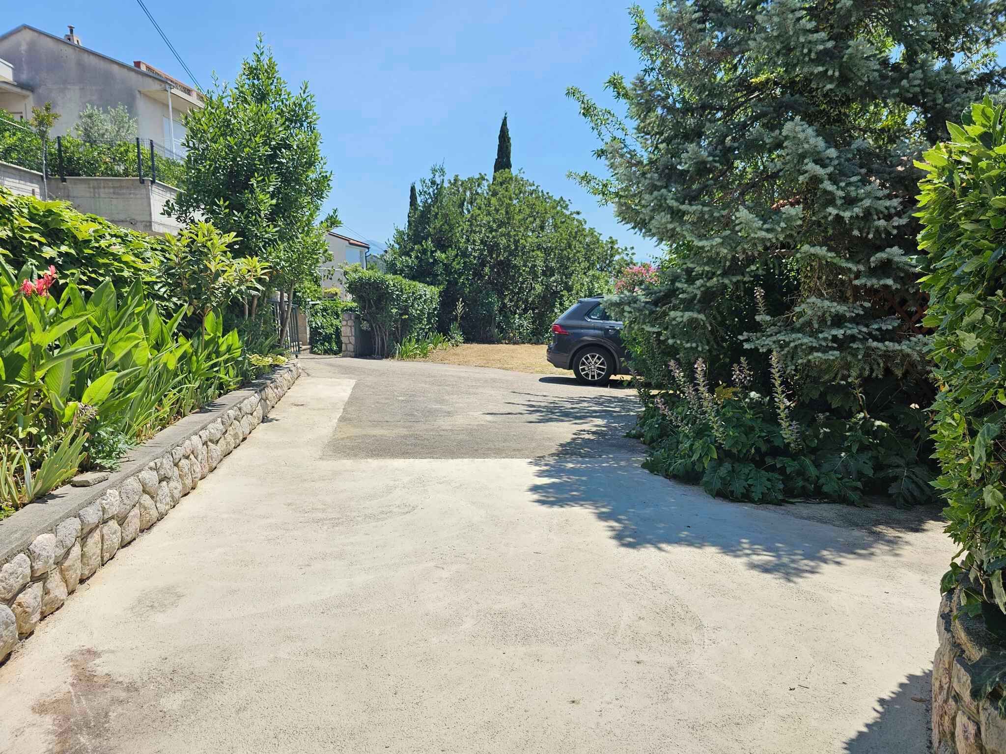 A residential street with a parked car, surrounded by lush greenery and a stone wall.