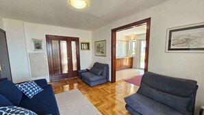 Uma sala de estar ampla com piso de madeira, dois sofás cinzas, um tapete estampado e uma cozinha visível por uma porta aberta.