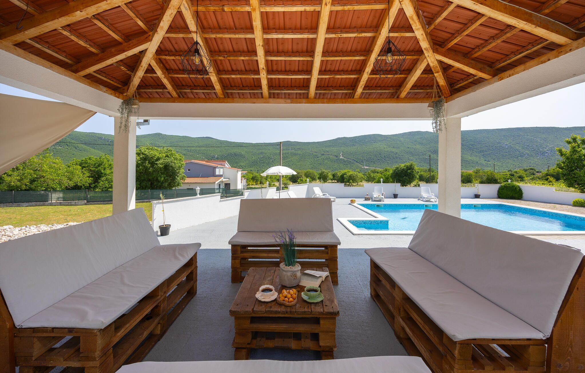 A covered outdoor seating area with wooden furniture, a pool, and a view of the mountains.