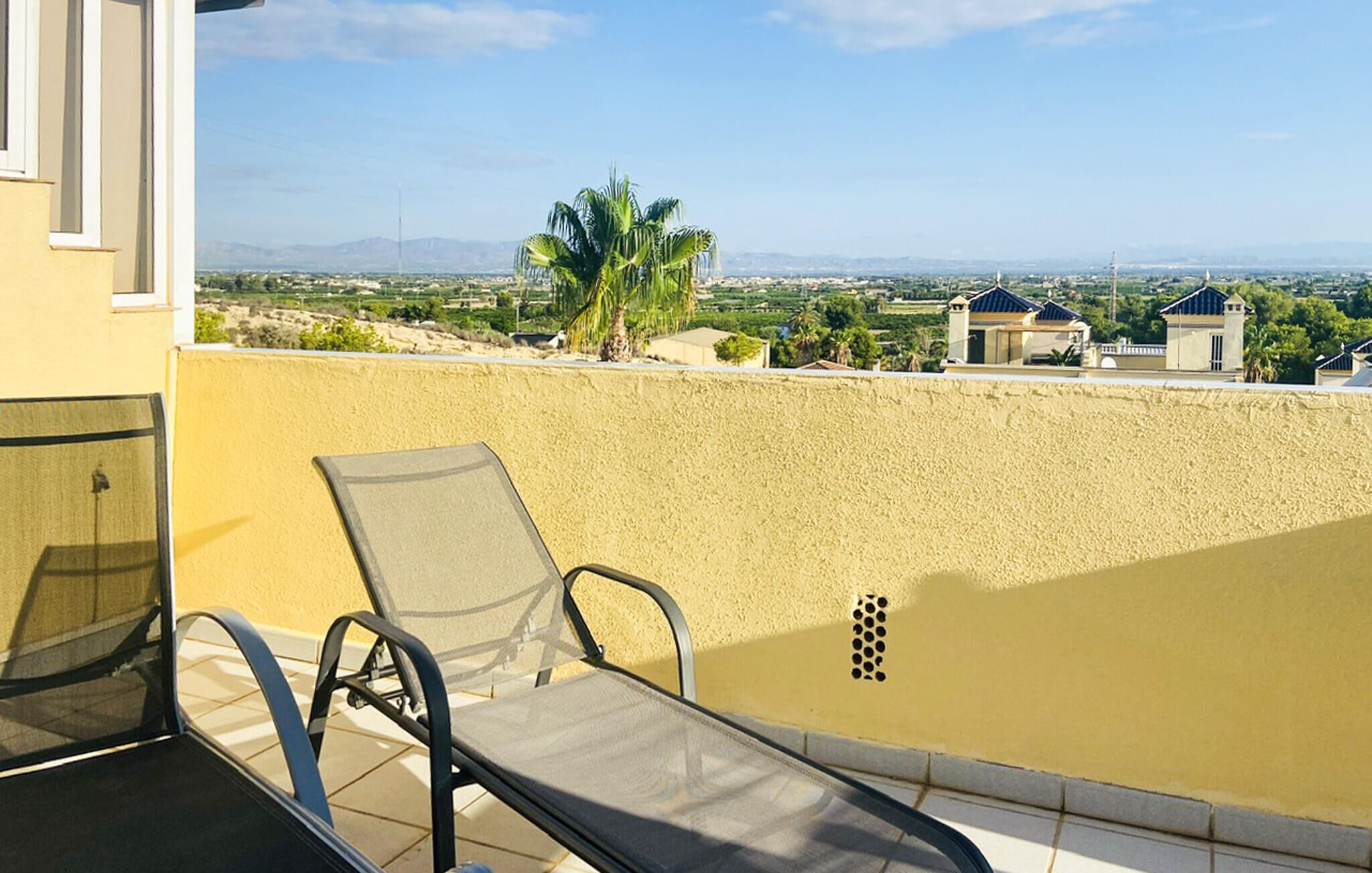 Un balcón con una silla y una mesa, con vistas a una zona residencial con casas y palmeras.