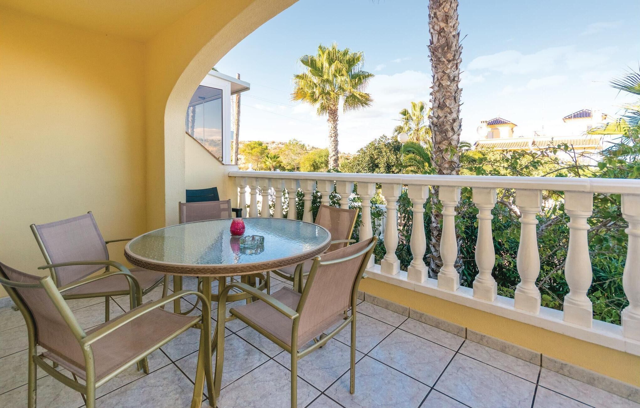 A balcony with a glass table and chairs, a railing, and a view of palm trees and buildings.