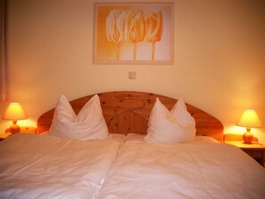 Uma cama de casal com duas almofadas, cabeceira de madeira, duas luminárias de cabeceira e um quadro de flores na parede.