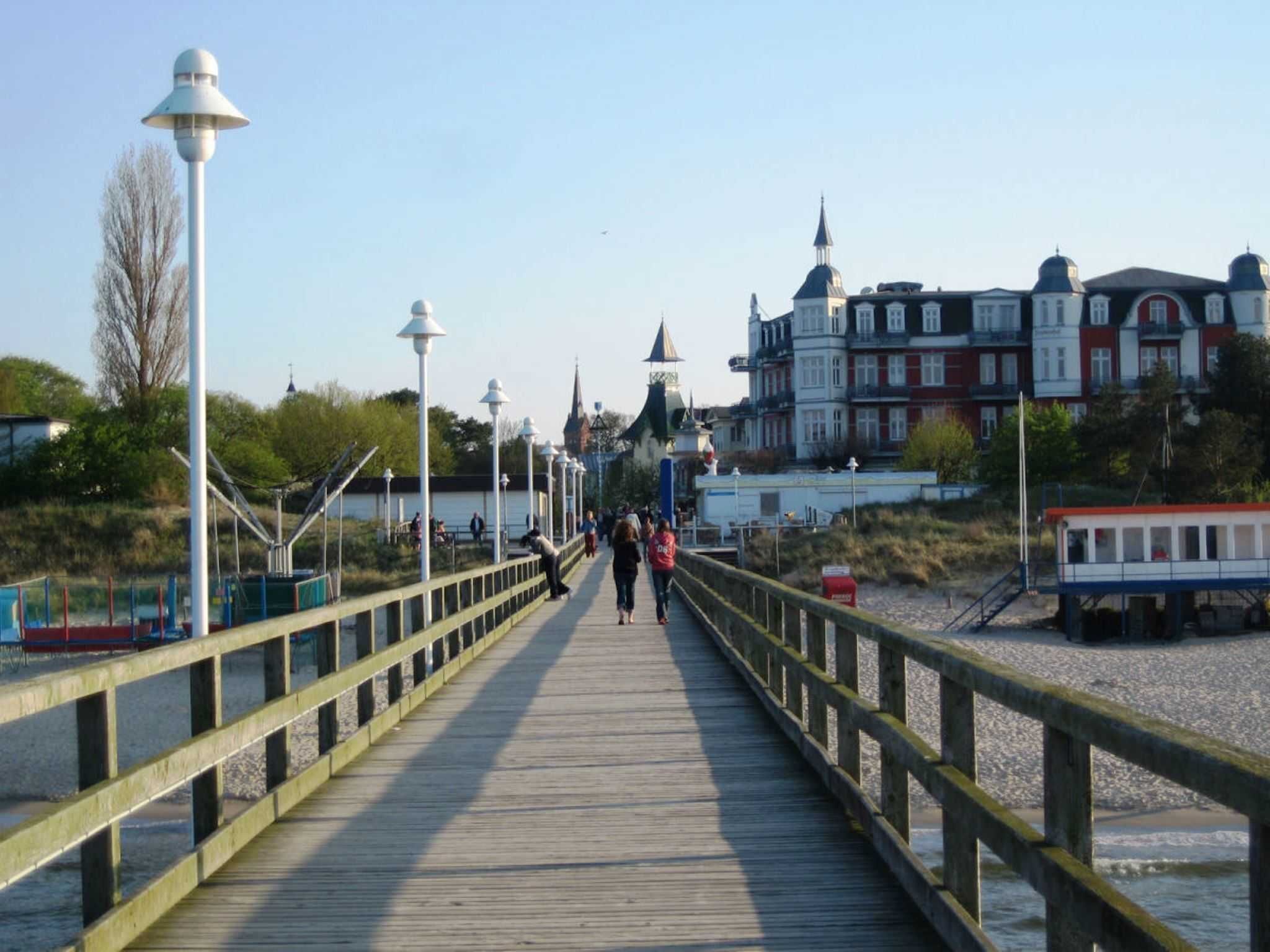 Una pasarela de madera con barandillas que conduce a una zona frente al agua, con edificios y un pequeño muelle.