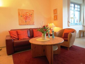 Uma sala de estar com um sofá marrom, uma mesa de centro redonda de madeira com um vaso de flores e uma cadeira de vime com almofadas laranja.