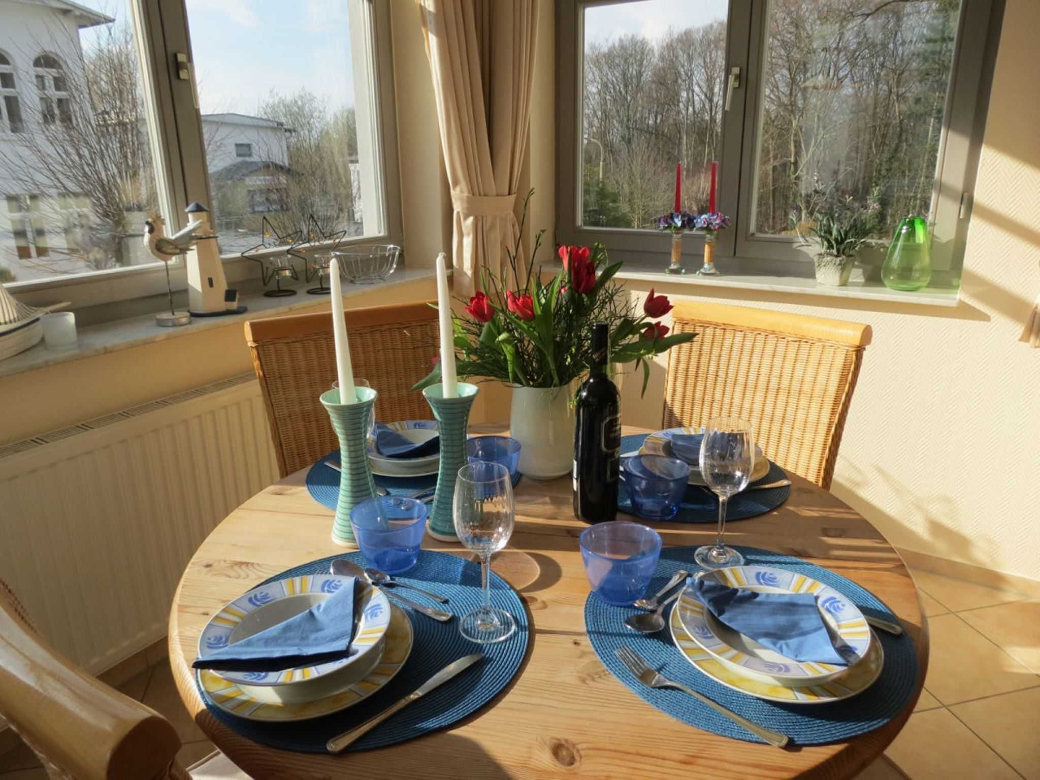 Une table à manger dressée avec des assiettes, des couverts et des verres, agrémentée d’un vase de fleurs et de bougies posés sur le rebord de la fenêtre.