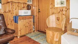 Une pièce aux murs lambrés, avec un fauteuil en cuir marron, un tabouret en bois orné d’un motif en forme de cœur, un micro-ondes blanc et un petit meuble avec des étagères.
