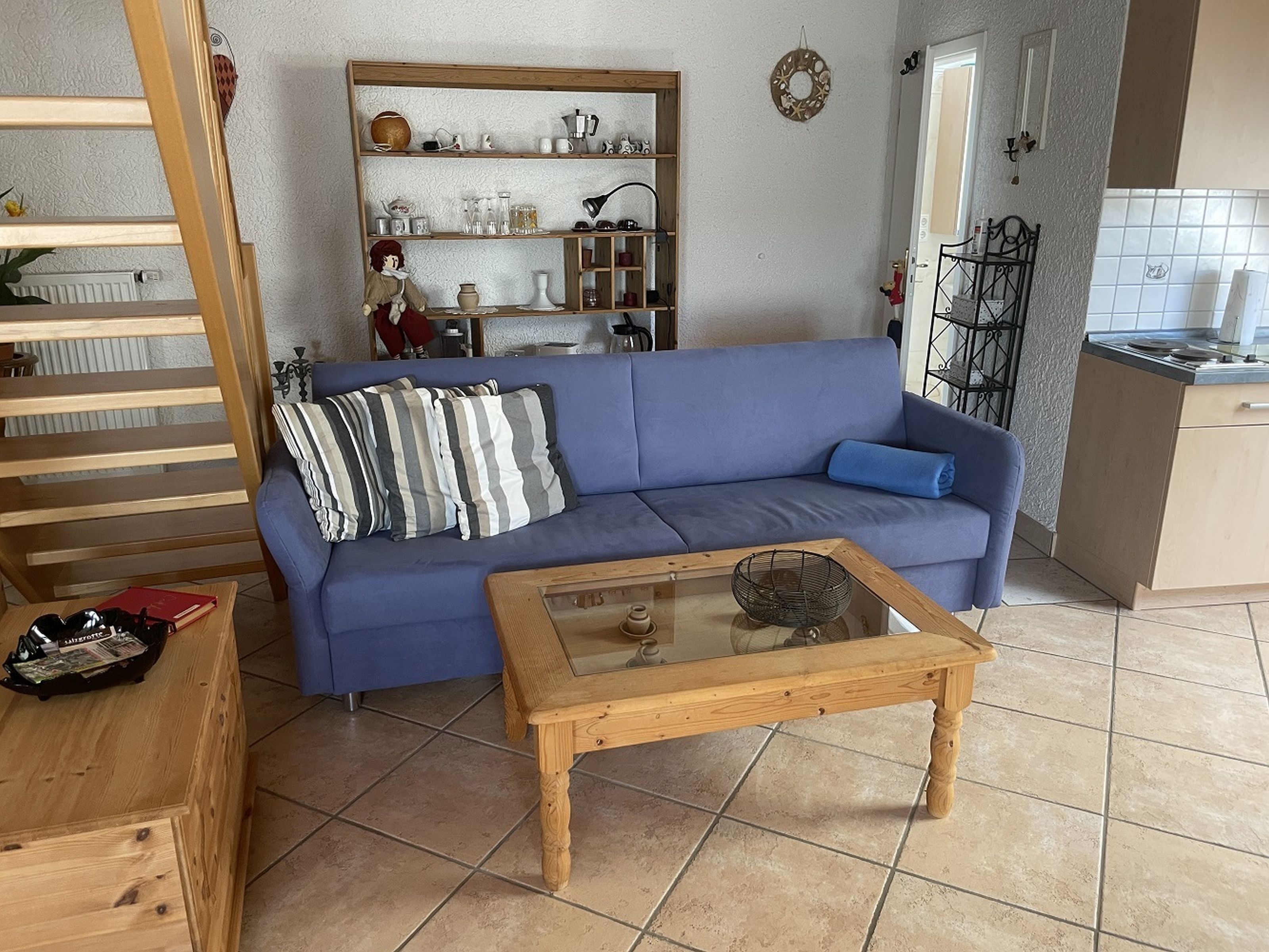 Ein Wohnzimmer mit blauem Sofa, einem Holztisch und einer Küchenzeile im Hintergrund.