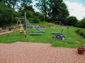 A playground with a slide, swings, and sun loungers on a grassy area with a brick pathway.