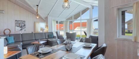 A spacious living area with a wooden ceiling, a large sofa, a dining table set with plates and glasses, and a view of a red-roofed building outside.