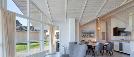 A spacious living area with a large window, a dining table with chairs, and a kitchen area in the background.
