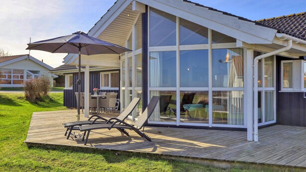Una casa moderna con un amplio ventanal de vidrio, una terraza de madera y mobiliario exterior.
