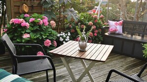 Une terrasse en bois avec une table et des chaises, entourée d’une végétation luxuriante et de plantes en fleurs.