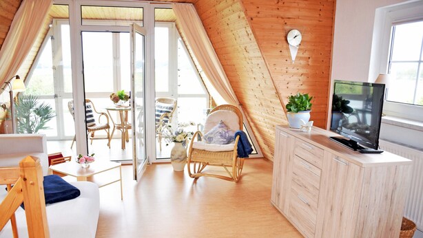 Une pièce chaleureuse avec un plafond en pente, un coin repas avec des chaises et une table, un fauteuil en osier, un téléviseur posé sur un meuble et une fenêtre avec des rideaux.