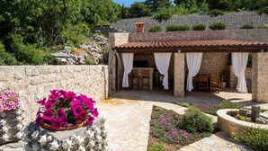 A stone-walled outdoor seating area with a tiled roof, surrounded by greenery and flowers. - 6 bedroom accommodation in Klek (Klek)