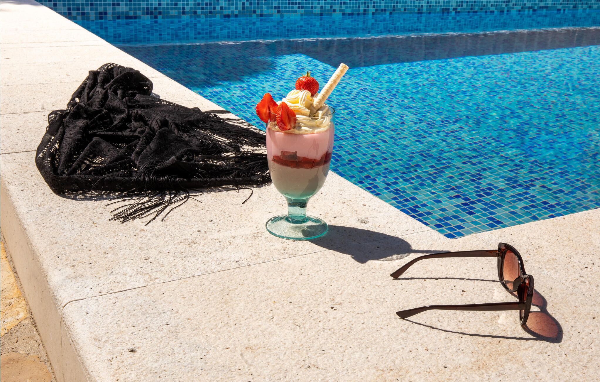 A glass of layered dessert with strawberries and whipped cream next to a pair of sunglasses, by a pool with blue tiles.