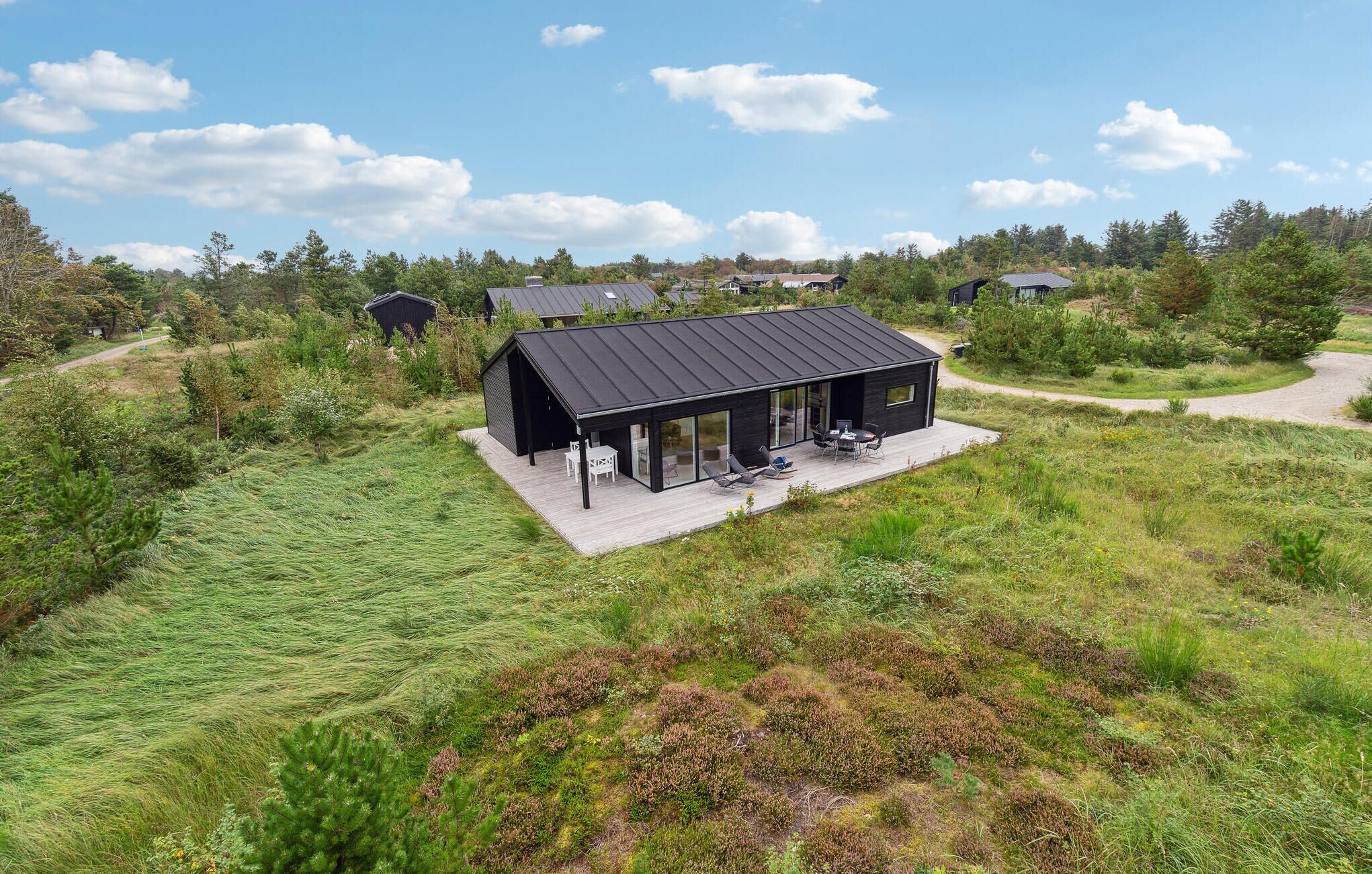 Una casa moderna negra con terraza, rodeada de paisaje natural y otras viviendas.