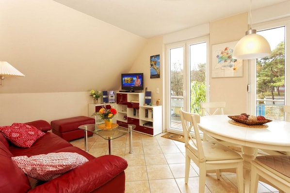 Un salon moderne avec un canapé rouge, une table basse en verre, une table à manger blanche avec des chaises, un téléviseur à écran plat et une grande fenêtre donnant sur la forêt.
