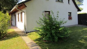 A single-story house with a sloped roof, a front porch, and a small garden area with a bush and flowers. - Vacation home Lorenz - Apartment 1/EG (Mölschow)