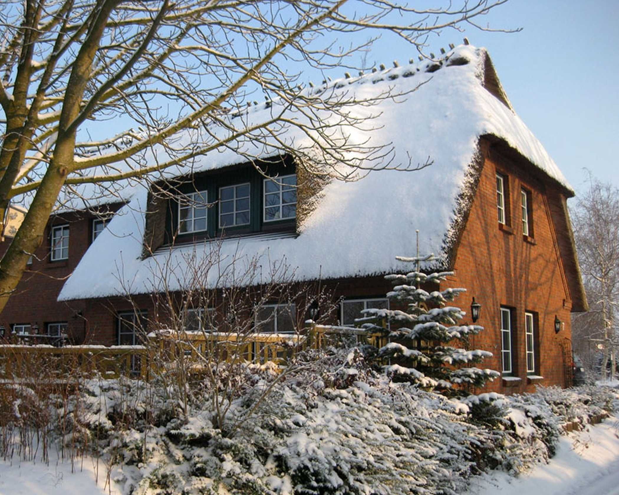 A traditional brick house with a steeply pitched roof covered in snow, surrounded by snow-covered bushes and trees.