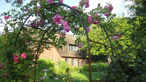 En have med et rosebuegang, et stråtækt hus og forskellige planter.