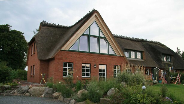 Et traditionelt hus med et stejlt stråtækt tag, store vinduer og en murstensfacade.