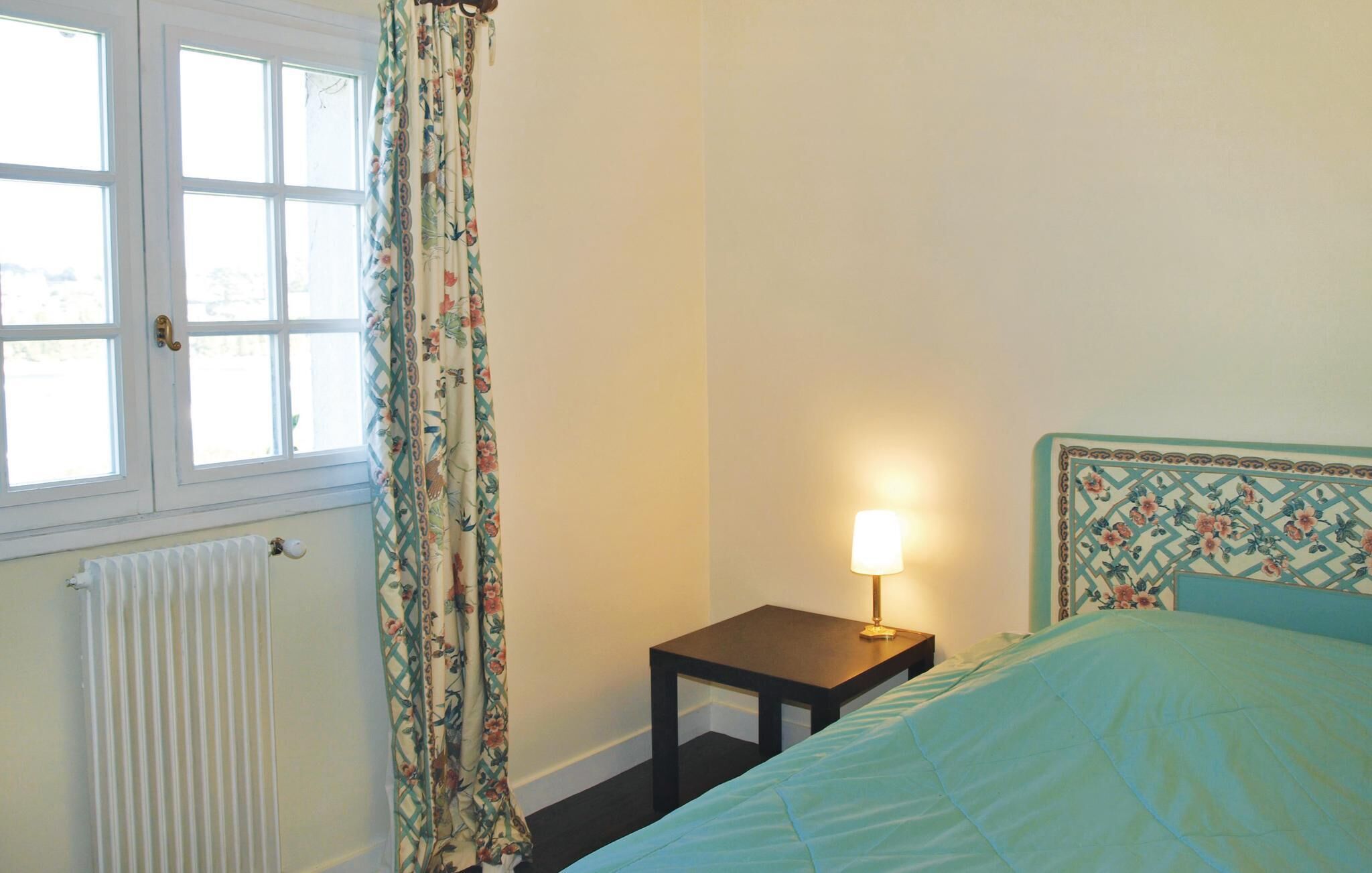 A bedroom with a bed, a bedside table with a lamp, a radiator, and a window with curtains.