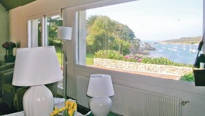 Une pièce dotée d’une grande fenêtre donnant sur un paysage côtier magnifique, d’une table avec une lampe et d’un vase rempli de fleurs.